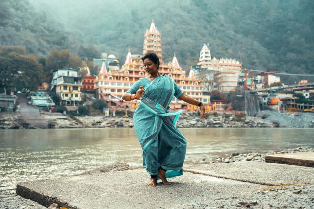 A woman in a turquoise sari dancing on a riverbank in front of a temple complex in Rishikesh