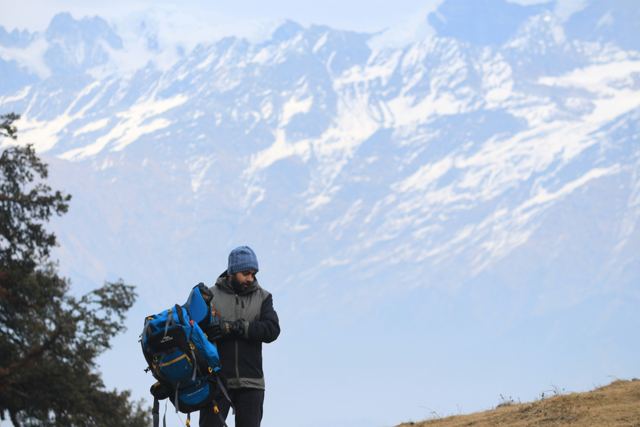 A trekker with a blue backpack on a grassy ridge facing a snow range